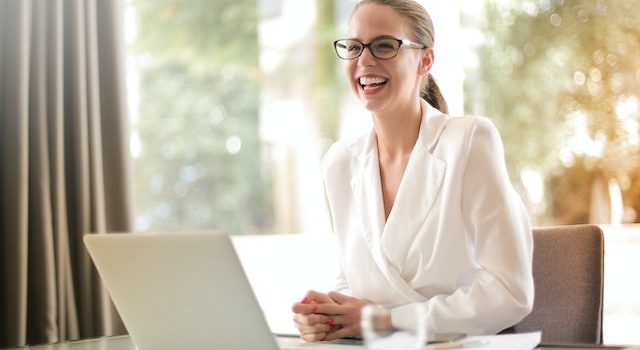 Wanita mengenakan kotoran putih duduk di depan laptop dan tersenyum