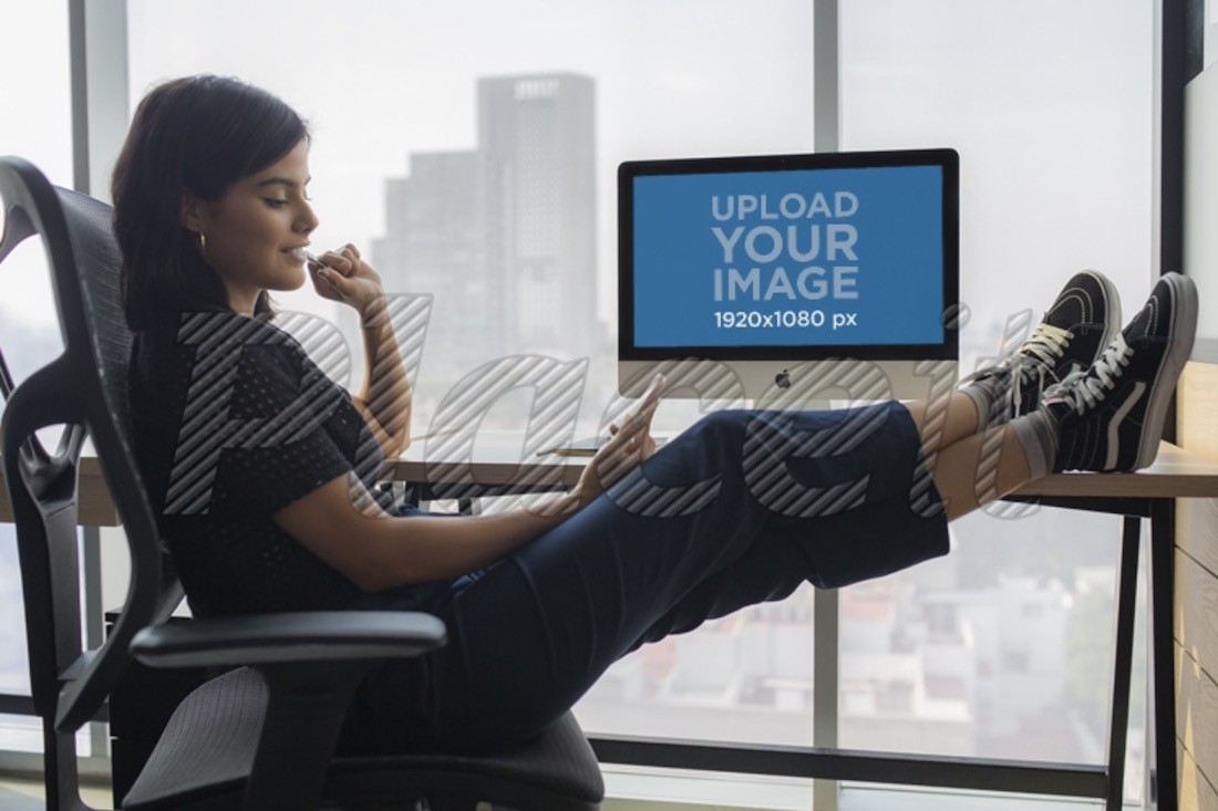 wanita memeriksa teleponnya di kantor dekat mockup iMac